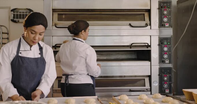 Female bakers exchanging cue in bakery, baker in coat loading deck oven trays for baking buns
