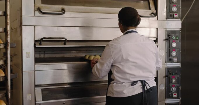 African American woman in apron opening oven in pink mitts, removing rolls onto rack for cooling