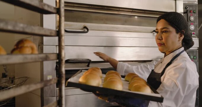Mid-adult woman baker in bakery opening oven, sliding tray of rolls into rack and adjusting spacing