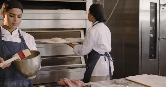 Two female bakers after proofing shaping dough and brushing glaze for baking in bakery kitchen