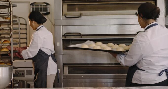 Diverse female bakers loading ovens after tray moving, pulling rack, checking roll inside at bakery