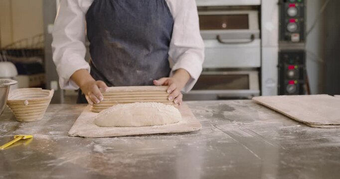 Middle-aged baker in apron at bench lifting basket showing dough shaping loaves on peel for baking