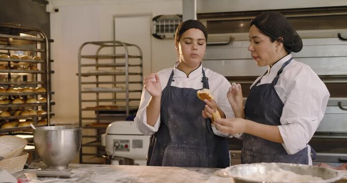 Diverse female coworkers in chef coats tasting rolls after left bite, checking quality on bench