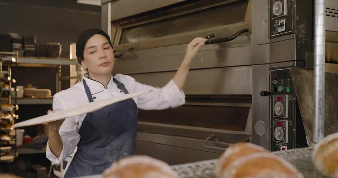 Two female bakers grabbing wooden peel placing tray into deck oven adjusting controls for baking