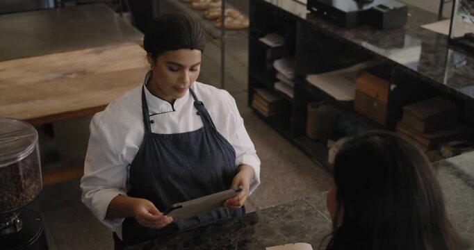 Female baker tapping tablet selecting pastry as customer with phone arriving to buy at counter