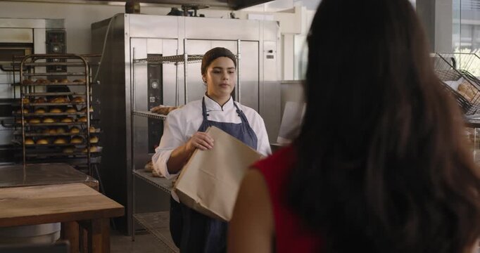 Female baker following female buyer request at counter taking bread in apron bagging, tapping card