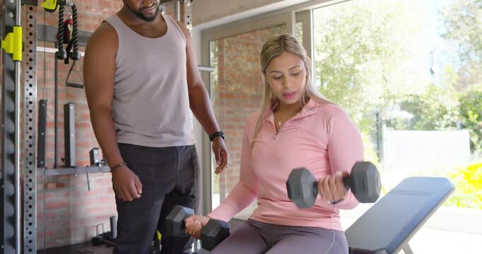 African American male trainer cueing female client curling hex dumbbells on bench to build strength