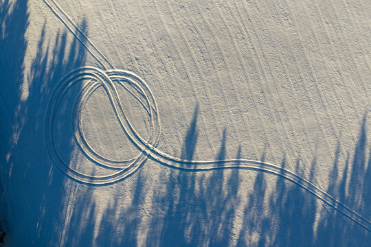 Aerial view of vehicle tracks forming circular patterns on a snow-covered field with long tree shadows in winter light Lithuania.