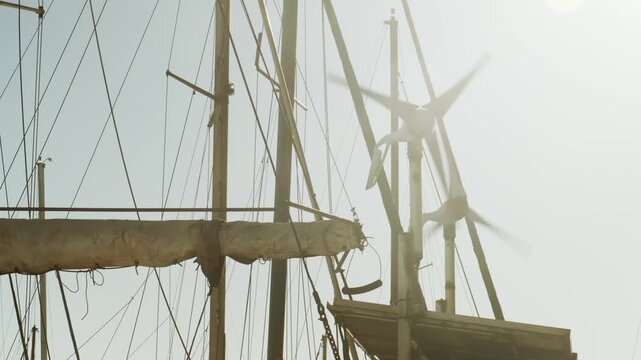 Tall ship masts and complex rigging silhouetted against bright hazy sky. Multiple ropes, spars and sails create dramatic nautical composition for maritime heritage content.