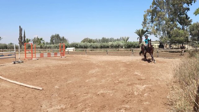 ​A dynamic, ground-level medium shot of a young equestrian rider, a teenager, cantering a powerful dark bay horse through a turn in a dirt training arena. 
