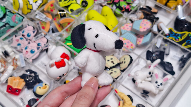 Close up of Hand Holding Snoopy Peanuts Jibbitz Charm for Crocs Shoes Customization Surrounded by Various Character Shoe Charms in Display Case on May 9, 2026 in Bangkok, Thailand 