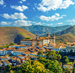 Panoramic view of the historic city of Ouro Preto, Minas Gerais, Brazil, featuring colonial architecture, Baroque churches, and terracotta roofs nestled against lush green mountains under a blue sky. © lcrribeiro33@gmail