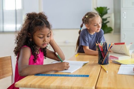 African American girl writing in notebook at desk with student classmate in classroom while studying at elementary school