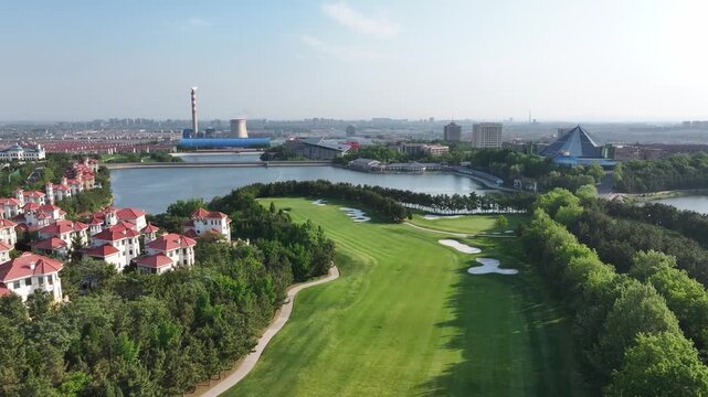 Golf course with lakeside villas and industrial area in the distance under clear summer sky
