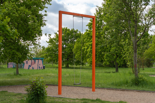 Orange swing set stands empty in grassy park. Trees surround area, sky bright with clouds. Quiet scene suggests calm, peaceful day. Ideal for advertising, travel, or childhood themes