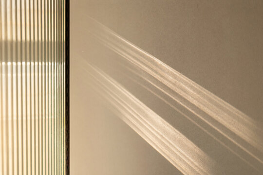 Abstract sunlight and shadow patterns cast through a fluted glass pane onto a neutral beige wall in a minimalist interior setting during golden hour for background use.