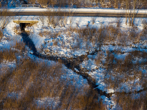 Aerial view of a small stream flowing from a culvert under a road through snow-covered reeds and winter vegetation Lithuania.