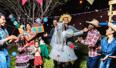 Naklejka premium Diverse Brazilian family and friends celebrating Festa Junina at night with traditional costumes, dancing, and a mock wedding ceremony in a festive outdoor setting.