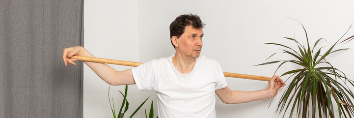 Obraz premium A 53-year-old man is working out at home. He is using a stick for balance and is focused on his exercise. The room features plants and a yoga mat on the floor, banner
