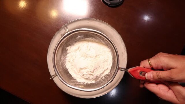 Hands Sifting white All purpose flour through a metal sieve into a bowl for baking