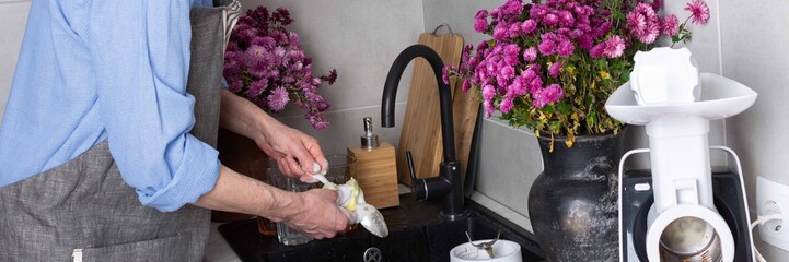 A man stands at a sink in a kitchen, washing dirty dishes and glassware. Flowers sit on the counter as fresh light comes in through the window. The setting is busy and practical, banner © pundapanda