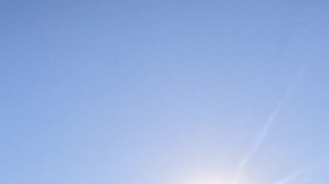 Large white seagull enters frame bottom left in slow motion, ascends and glides right across bright blue sky with sun glare, wings fully extended, then exits right edge of frame.