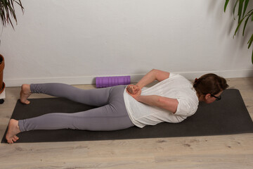 Naklejka premium A woman aged 52 stretches on a yoga mat at home. She is focusing on her workout while practicing a back stretch. Plants are visible in the background