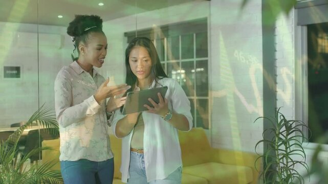 Two women leaning near sofa, right woman tapping tablet and animating charts for marketing review