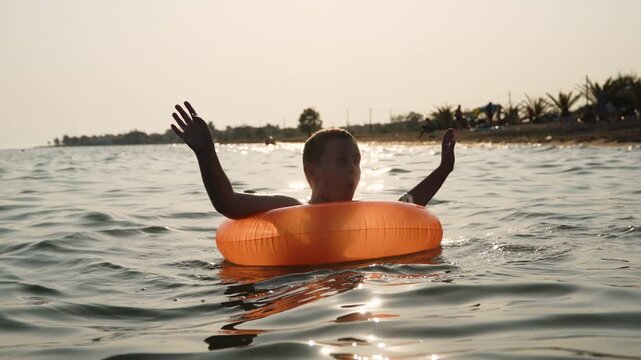 Caucasian boy happily spins in inflatable ring in sea at sunset. Enjoyment and euphoria of child from swimming in water during travel. Water therapy in warm climate.