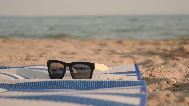 Sunglasses lie on woven beach mat on golden sand with sea waves closeup. Aesthetic background shot of beach vacation and UV protection.