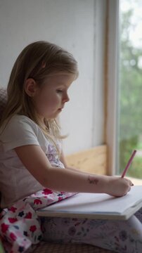 Focused, sweet preschool girl drawing with pencils on paper at home by the window, engaging in creative activities, developing her creativity and imagination, developing skills, and enjoying art.