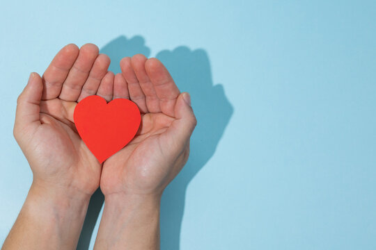 Male hands together holding a paper heart on a light blue background with copy space. Concept of care, support, well being, emotional health, and health protection