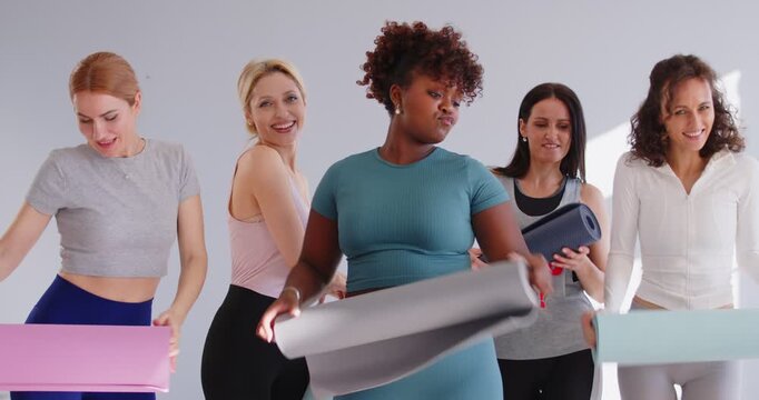Happy sporty diverse women group having fun in gym with yoga mat in hands and looking cheerful at camera after fit training. Sport workout and healthy lifestyle concept. 4k video. Slow motion video.
