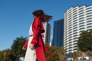 Obraz premium A woman in a red trench coat walks outdoors near modern buildings on a sunny day. Her hair flows in the wind, capturing movement and urban lifestyle.