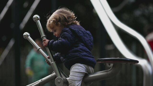 Toddler climbing onto playground equipment gripping metal bars with focused effort showing early motor skills determination cautious movement and natural childhood exploration in park