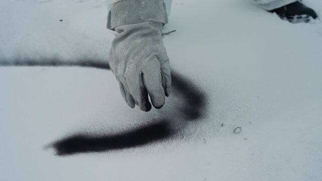 Hand sprinkles black powder on snow to create Sshaped trail, gloved technician draws sweeping curve, fine grains scatter across pristine surface, boot prints nearby, closeup contrast between dark