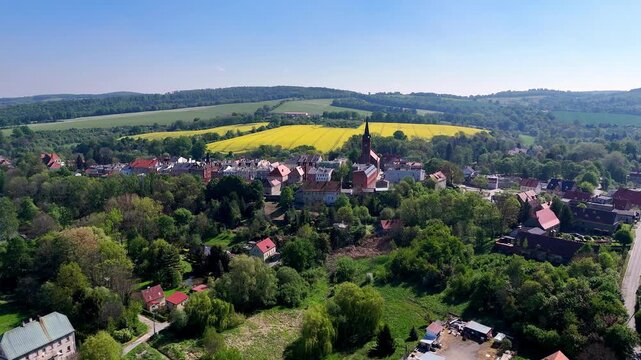Scenic aerial movieof the historic town of Niemcza, Poland, featuring a prominent church tower, colorful townhouses, and red rooftops, surrounded by lush green trees and yellow rapeseed fields under a
