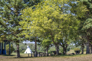 Beautiful summer park scene in the summer with lush, green foliage, wooden picnic table under the shade and a tipi in the background. © Jolanta