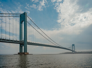 Fototapeta premium Verrazzano Narrows Bridge Over New York Harbor. Verrazzano-Narrows Bridge over harbor water with suspension cables, clouds, and hazy shoreline.