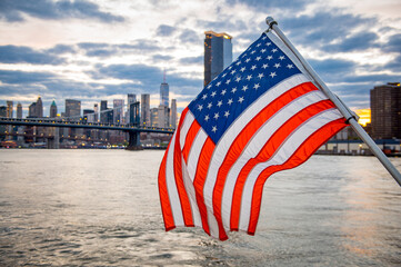 Naklejka premium American Flag Over East River And Manhattan Skyline. American flag in foreground with East River, Brooklyn Bridge, and Manhattan skyline at sunset.