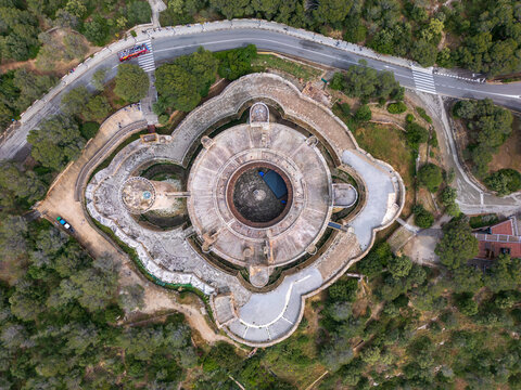Aerial view of the circular Castillo de Bellver with its unique Gothic architecture surrounded by lush green pine forests and a winding road in Palma, Illes Balears, Spain.