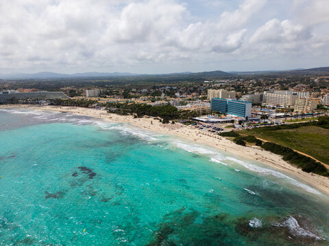 Aerial view of the turquoise waters and sandy beach with people relaxing near coastal hotels and resorts in Sa Coma, Balearic Islands, Spain.