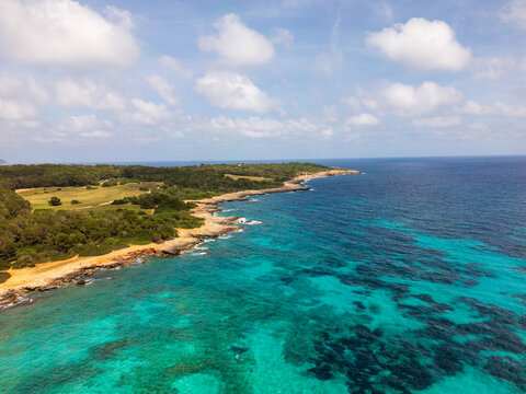 Aerial view of the rocky coastline and turquoise waters of the Mediterranean Sea under a blue sky with white clouds in Sa Coma, Balearic Islands, Spain.