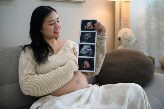 Smiling pregnant woman holding ultrasound photos while relaxing at home, representing pregnancy, prenatal care, and excitement for motherhood