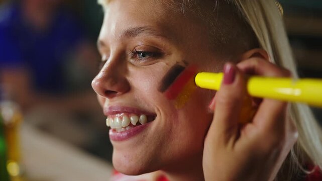 Close up of happy female football supporter getting German flag face paint from friend before watching sports match and cheering for national team.