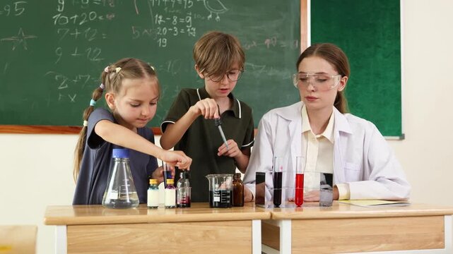 A female teacher wearing safety goggles guides two young students conducting colorful chemistry experiments in a classroom with a chalkboard full of math equations.