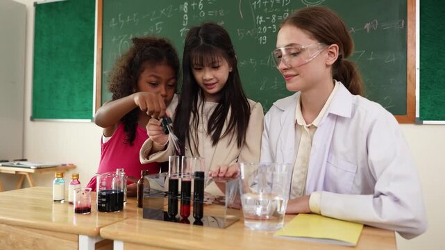 A female teacher and two girls conduct a chemistry experiment with test tubes and liquids in a classroom with a chalkboard in the background.