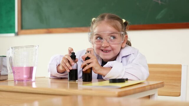 A young girl wearing safety goggles and a lab coat is surprised while examining two bottles in a science classroom.