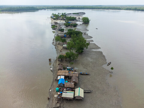 Aerial view of Kalabagi village on a narrow strip of land with small huts, lush trees, and wooden boats along the muddy riverbanks Khulna District, Khulna Division, Bangladesh.