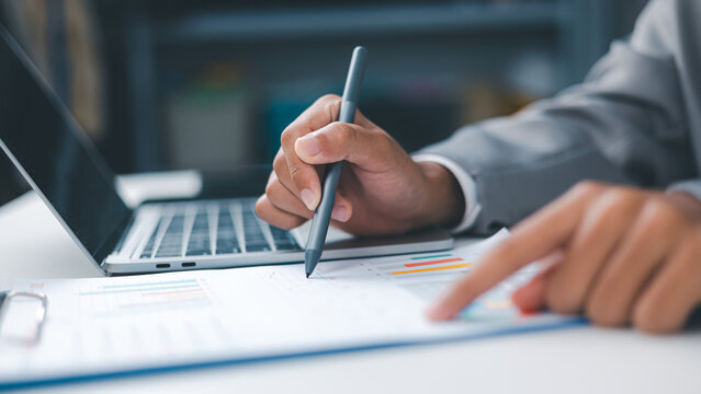 Businessman analyzing financial report charts and graphs on document with stylus pen and laptop. Concept of data analysis, auditing, investment planning, and corporate accounting technology.